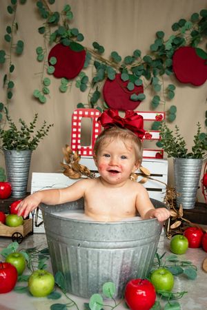A baby sits in a galvanized metal tub surrounded by red and green apples and decorative vines.