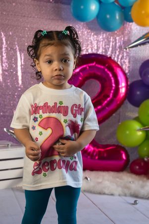 A child wearing a birthday-themed t-shirt stands in front of colorful balloons and party decorations.