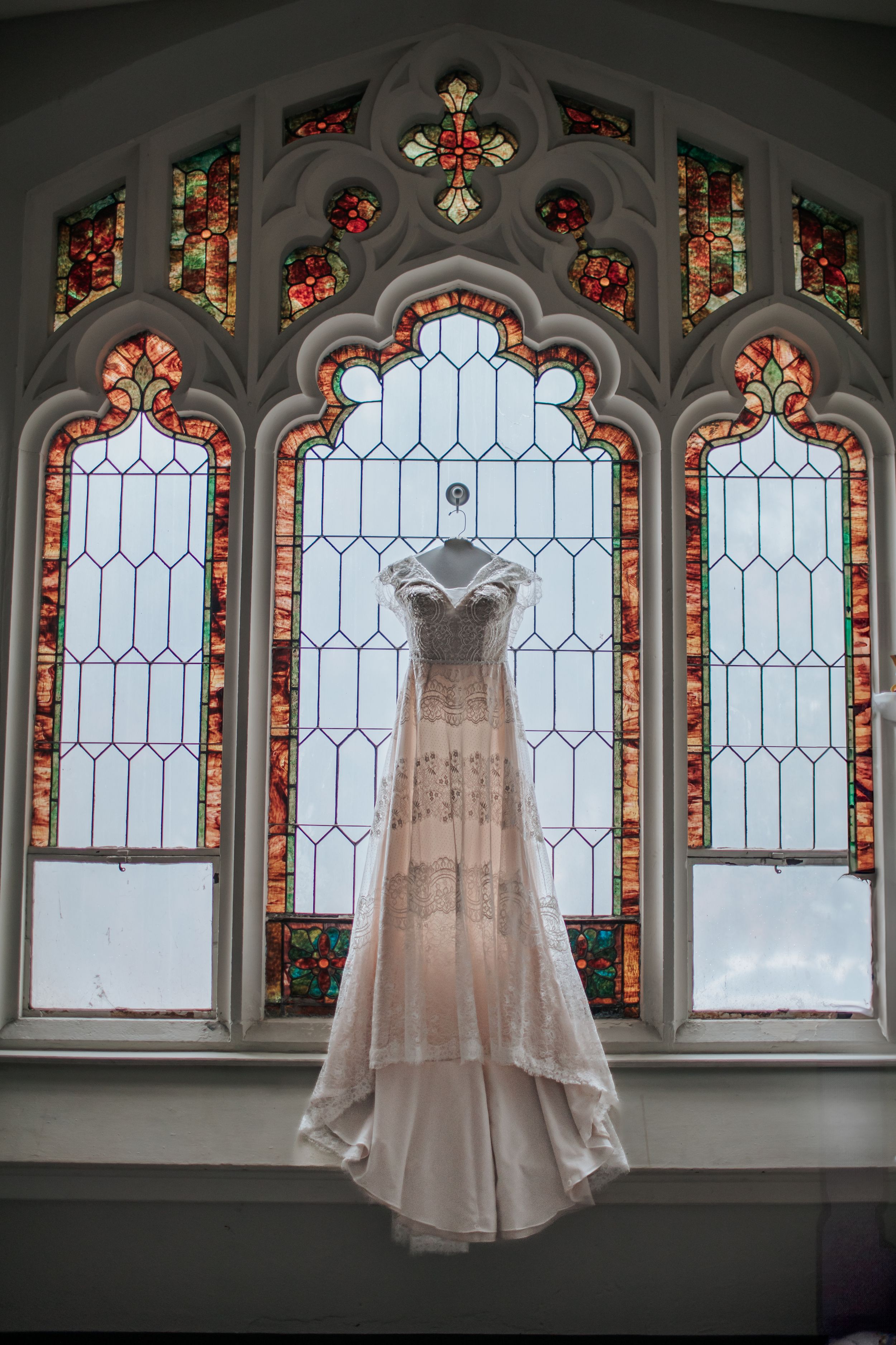 A white wedding dress hangs in front of ornate stained glass church windows with floral designs and gothic arches.