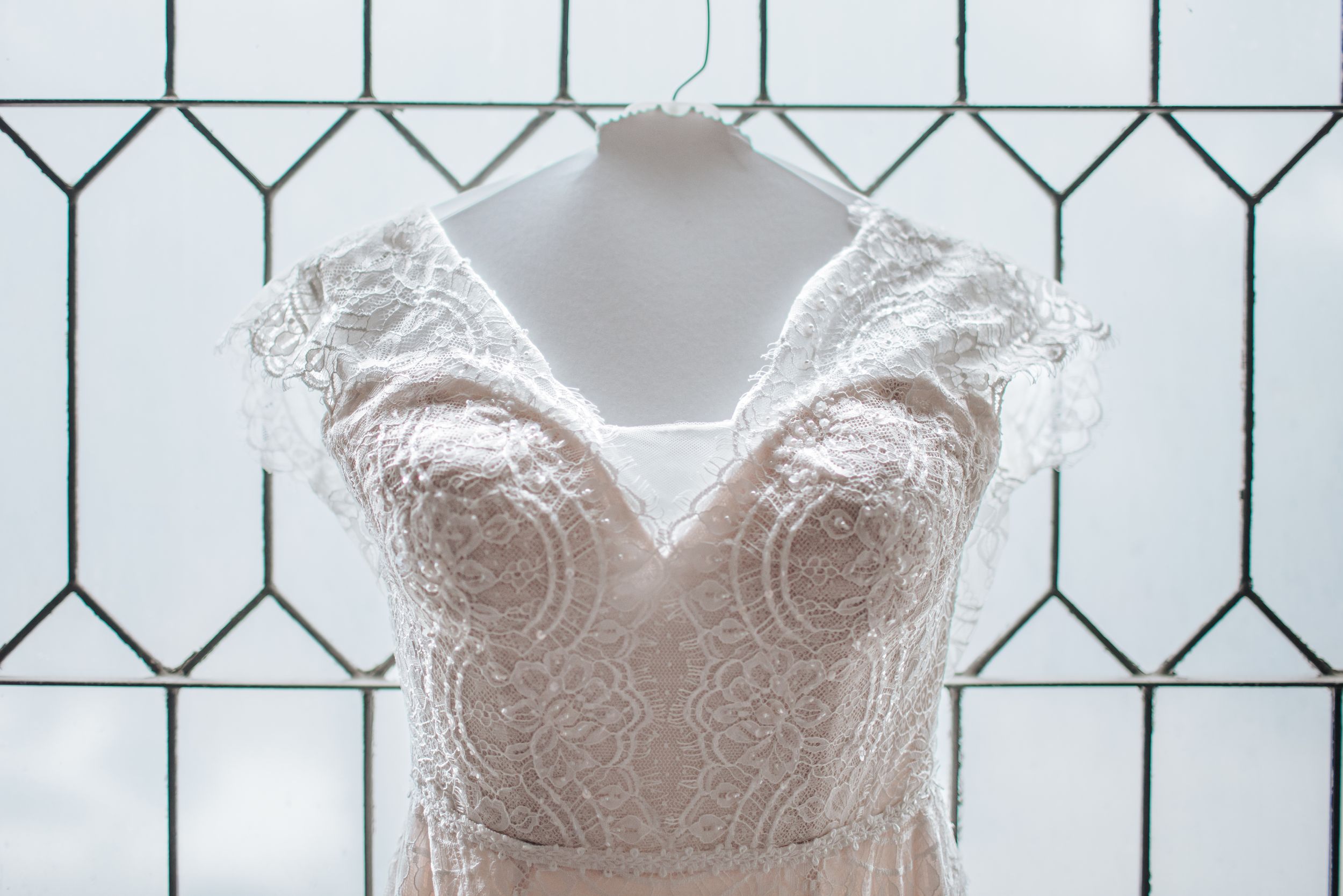 Wedding dresses displayed on hangers against a white wall with geometric pattern window panes.