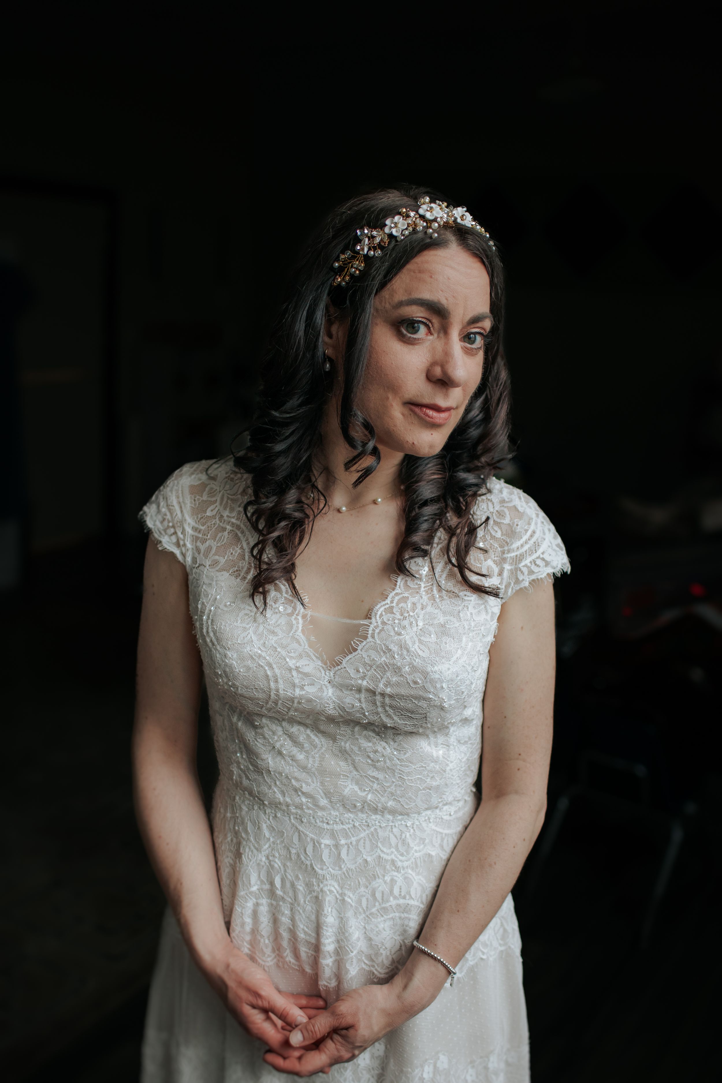 A bride in a delicate lace wedding dress with cap sleeves and a crystal headpiece poses in moody lighting.