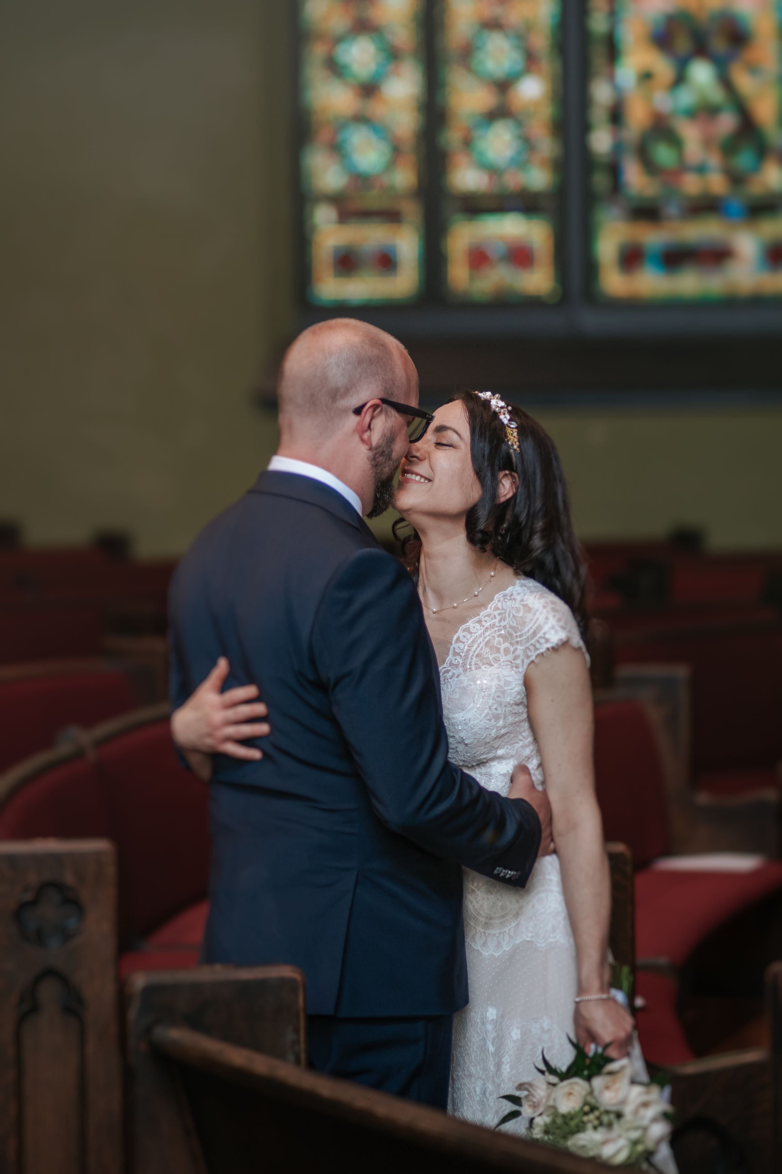A romantic wedding moment captured in a church with stained glass windows and wooden pews in the background.