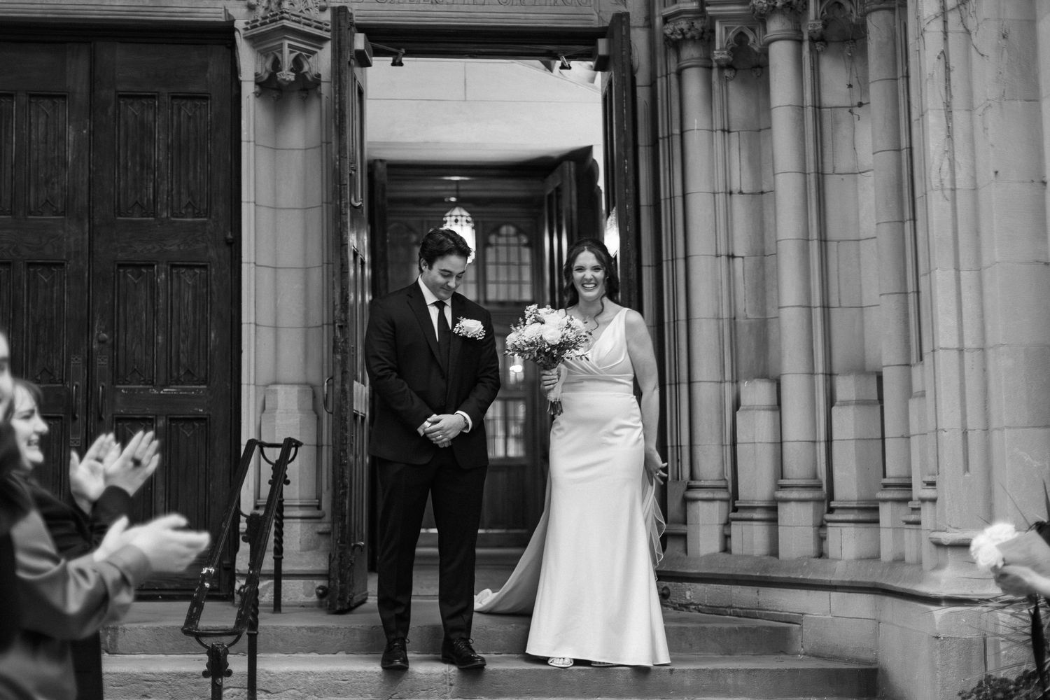 Wedding couple shares joyful moments with guests outside a stone church building in black and white photography.