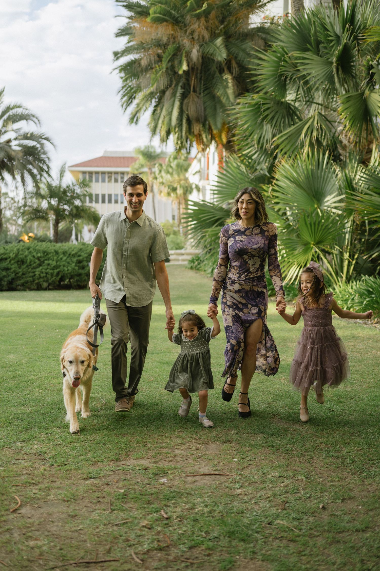 A group walks together holding hands under palm trees on a grassy lawn in coordinated brown and white outfits.