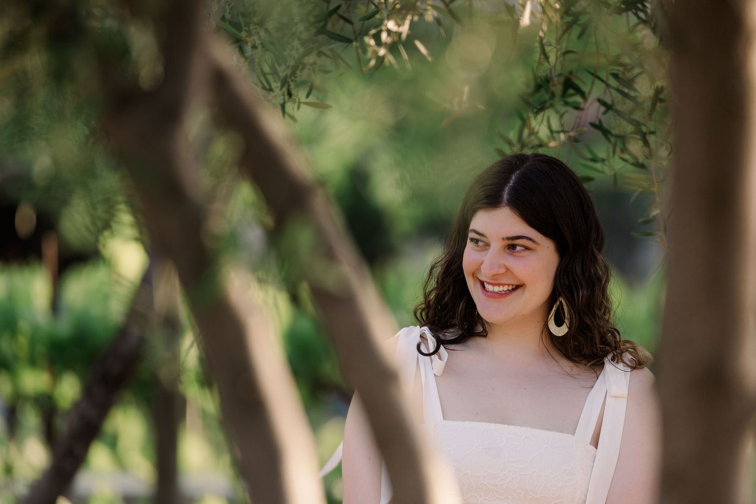 A person in a white dress smiles while posing in a garden setting with trees and natural light.
