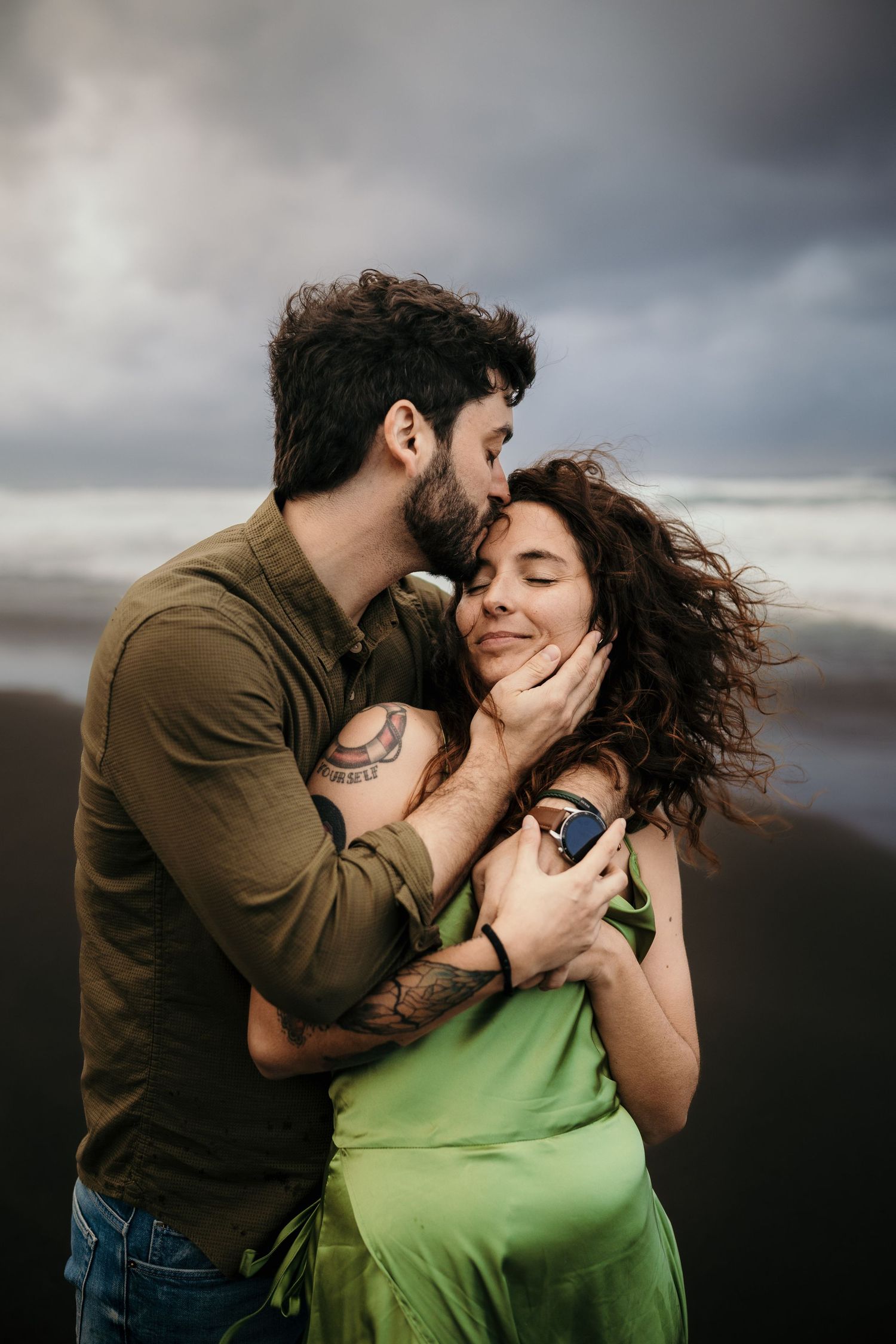 A couple embraces on a dark beach, man kissing woman's cheek as she smiles, her green dress flowing in the wind.