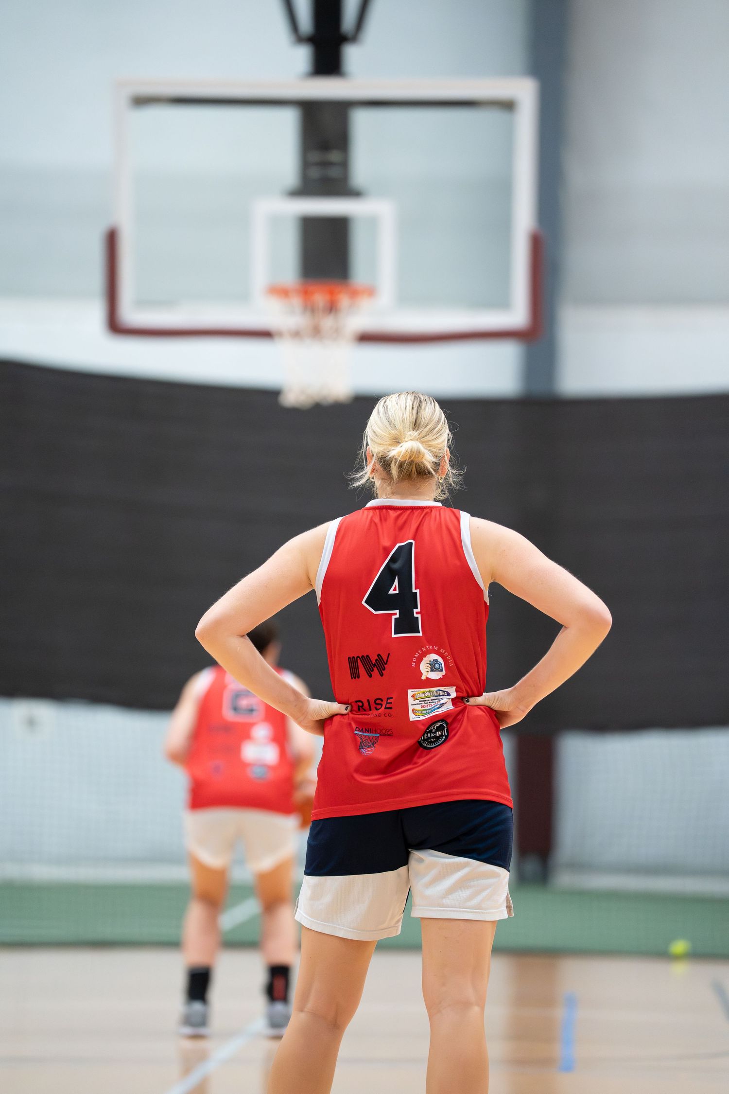 Female basketball player wearing red number 4 jersey stands on indoor court with basketball hoop in background.
