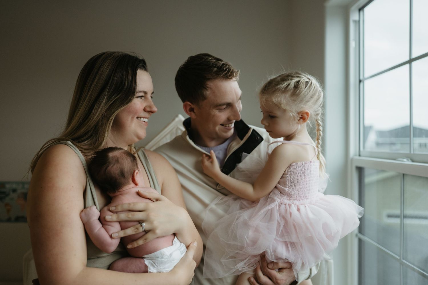 A happy family of four poses by a window, parents holding a newborn and toddler girl in a pink tutu dress.