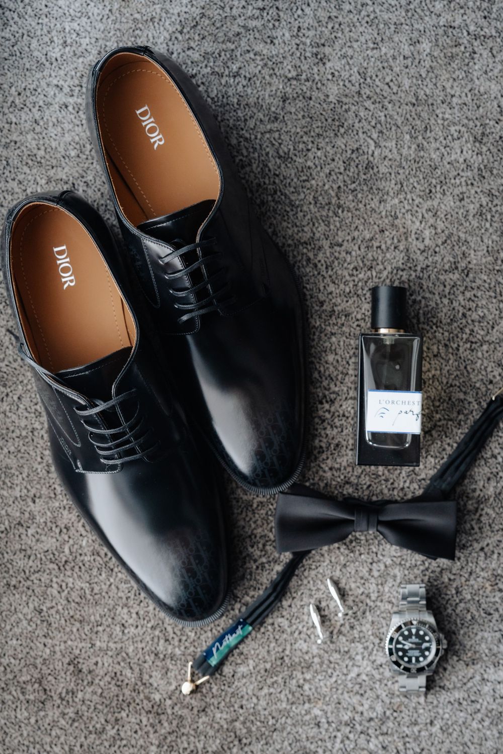 Black Dior dress shoes displayed with a bow tie, Rolex watch, cufflinks, and cologne on gray carpet.