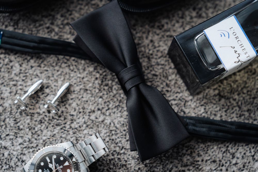 Black bow tie, silver cufflinks, and a classic wristwatch arranged on gray carpet beside a cologne box.