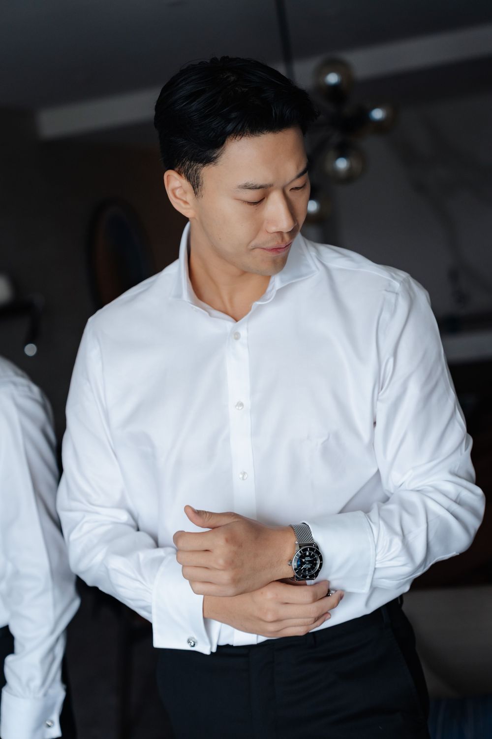 Asian man in white dress shirt adjusting cufflinks with a watch on his wrist, getting dressed formally.