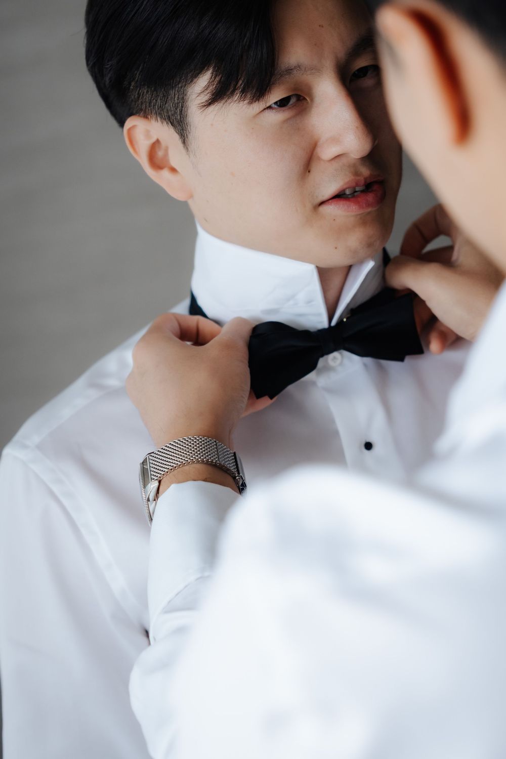 A man in a white dress shirt having his black bow tie adjusted by another person wearing a watch.