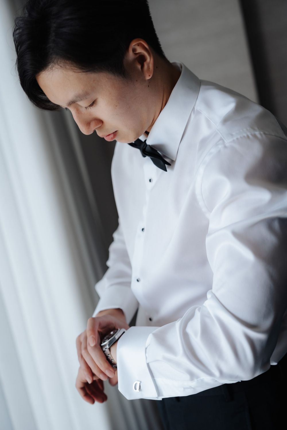 Elegant groom in white tuxedo shirt and black bow tie fastening his cufflinks.