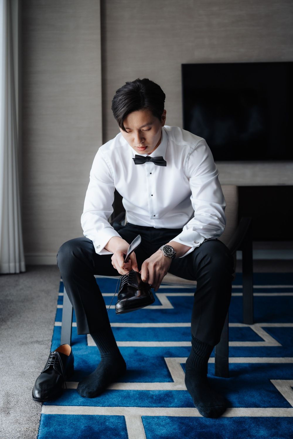 Asian groom in white tuxedo shirt and bow tie putting on black dress shoes while seated on blue geometric rug.