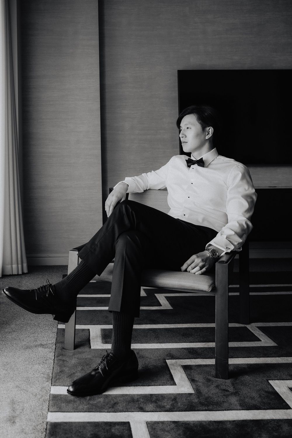 Elegant man in white tuxedo shirt and bow tie sitting relaxed in chair, black and white portrait photo.