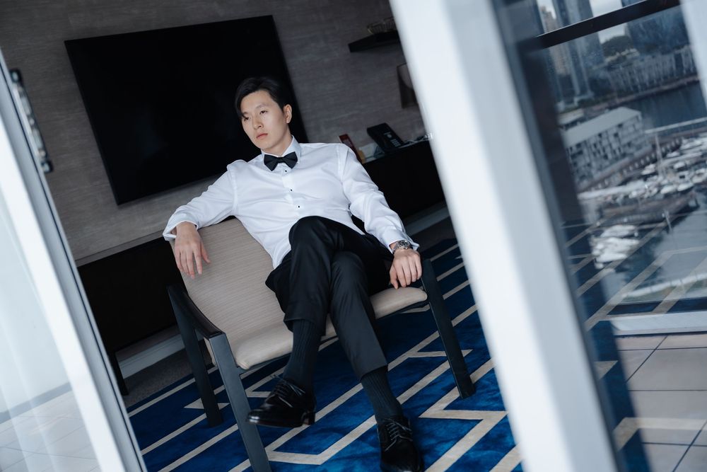 Elegant man in tuxedo sitting relaxed in modern hotel room with city skyline view through large windows.