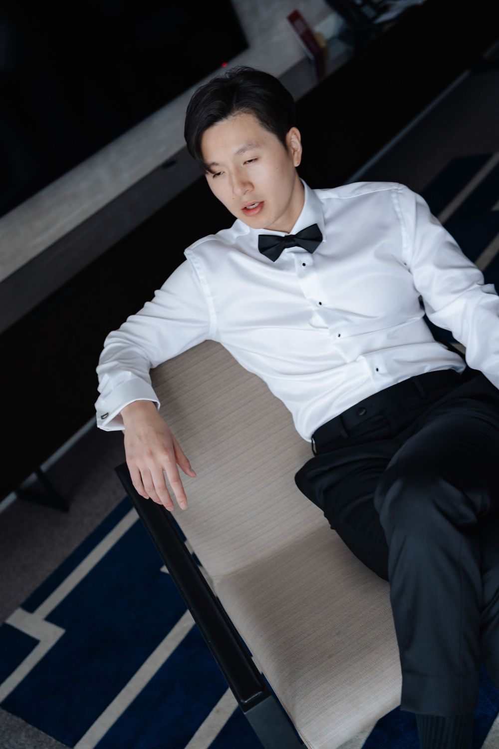 Young man in white dress shirt and black bow tie lounging casually in a chair at formal event.