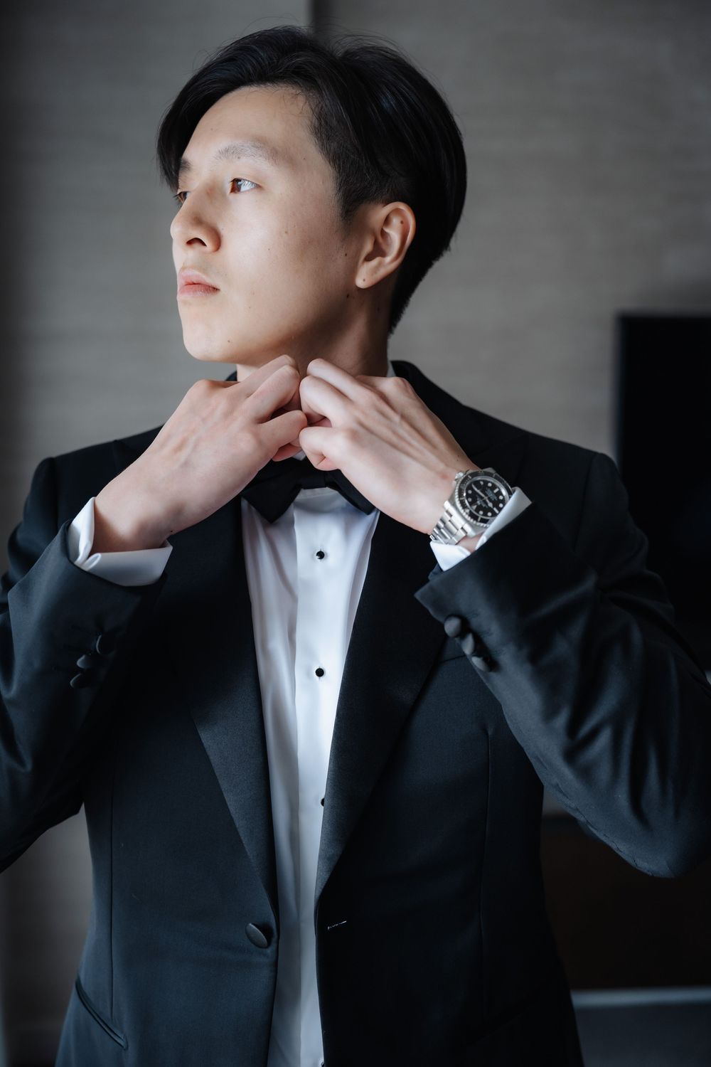 A young Asian man in a black tuxedo adjusts his bow tie, wearing a silver watch, in an elegant indoor setting.
