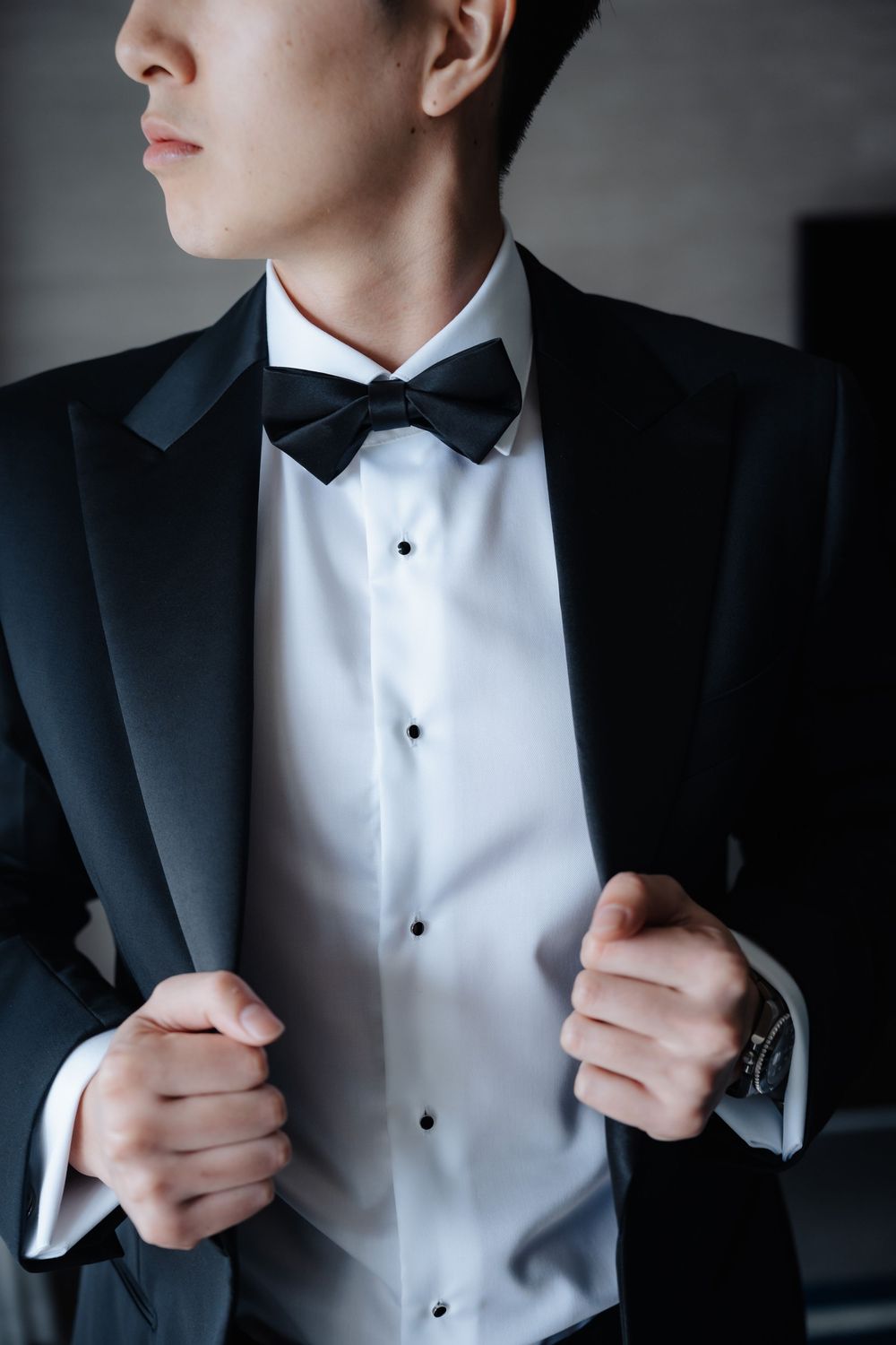 A man in a black tuxedo with bow tie adjusts his lapels, wearing a watch and formal white shirt.