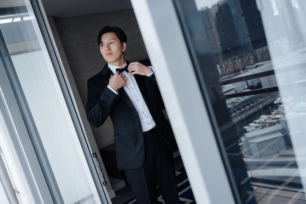 A man in a black tuxedo adjusts his bow tie while standing by floor-to-ceiling windows with a city view.