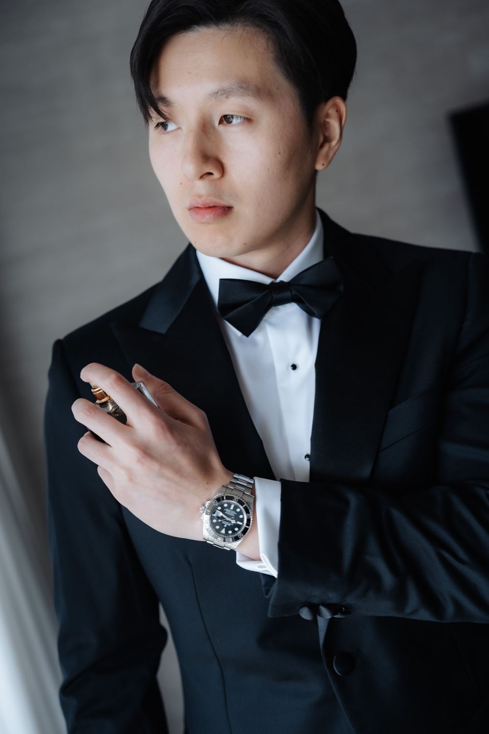 Young Asian man in black tuxedo and bow tie, wearing a silver watch and ring, posing elegantly indoors.