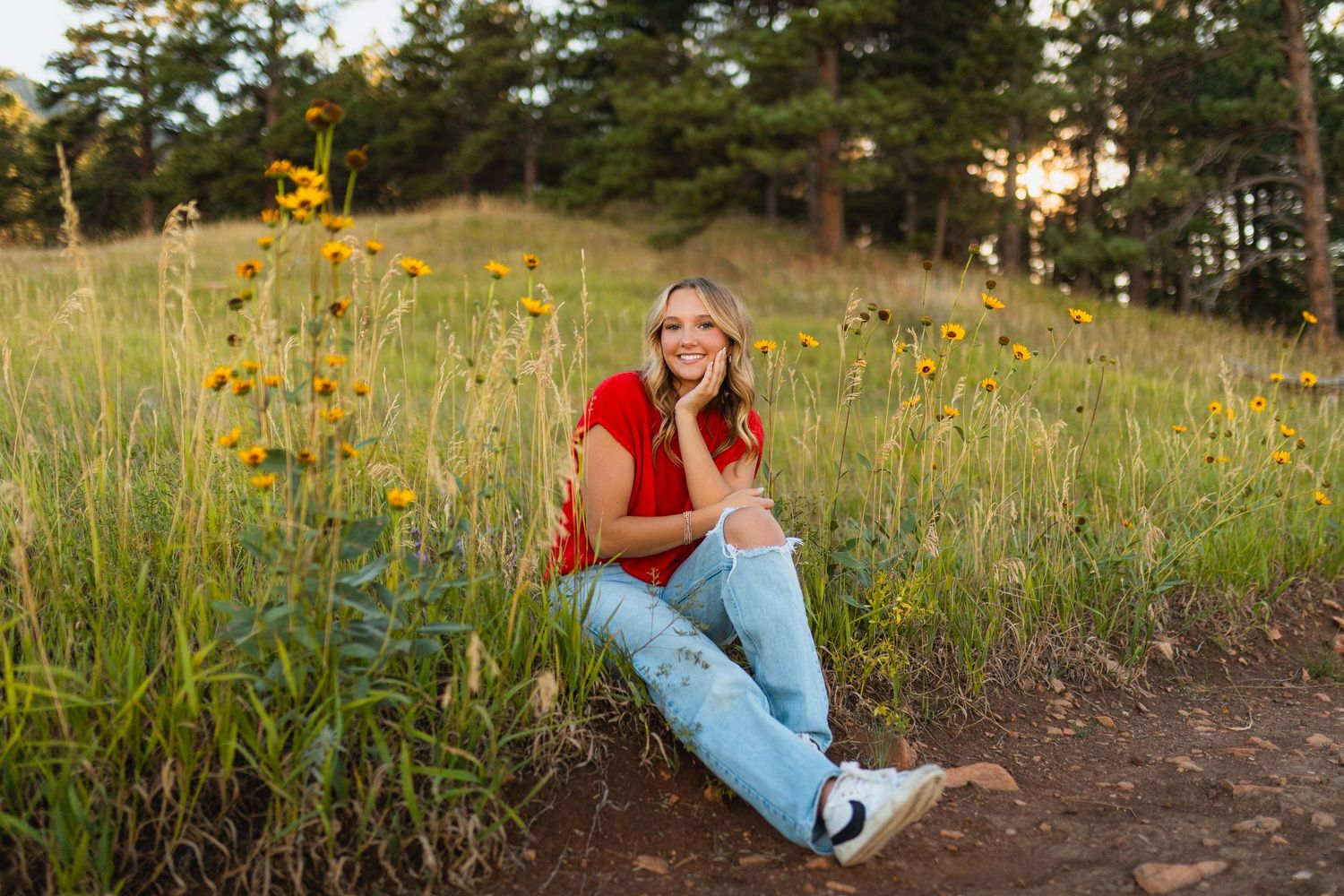 A smiling woman in a red top and light jeans sits among yellow wildflowers in a sunlit meadow with pine trees.