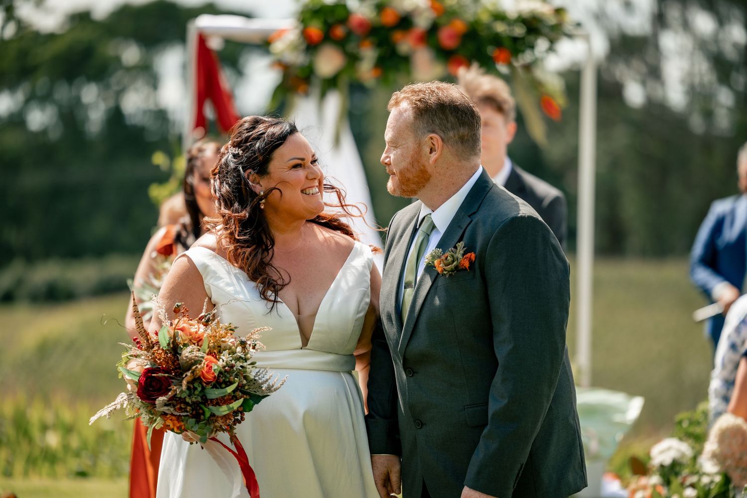 Bride and groom smiling at each other during outdoor wedding ceremony with floral arch and greenery backdrop.