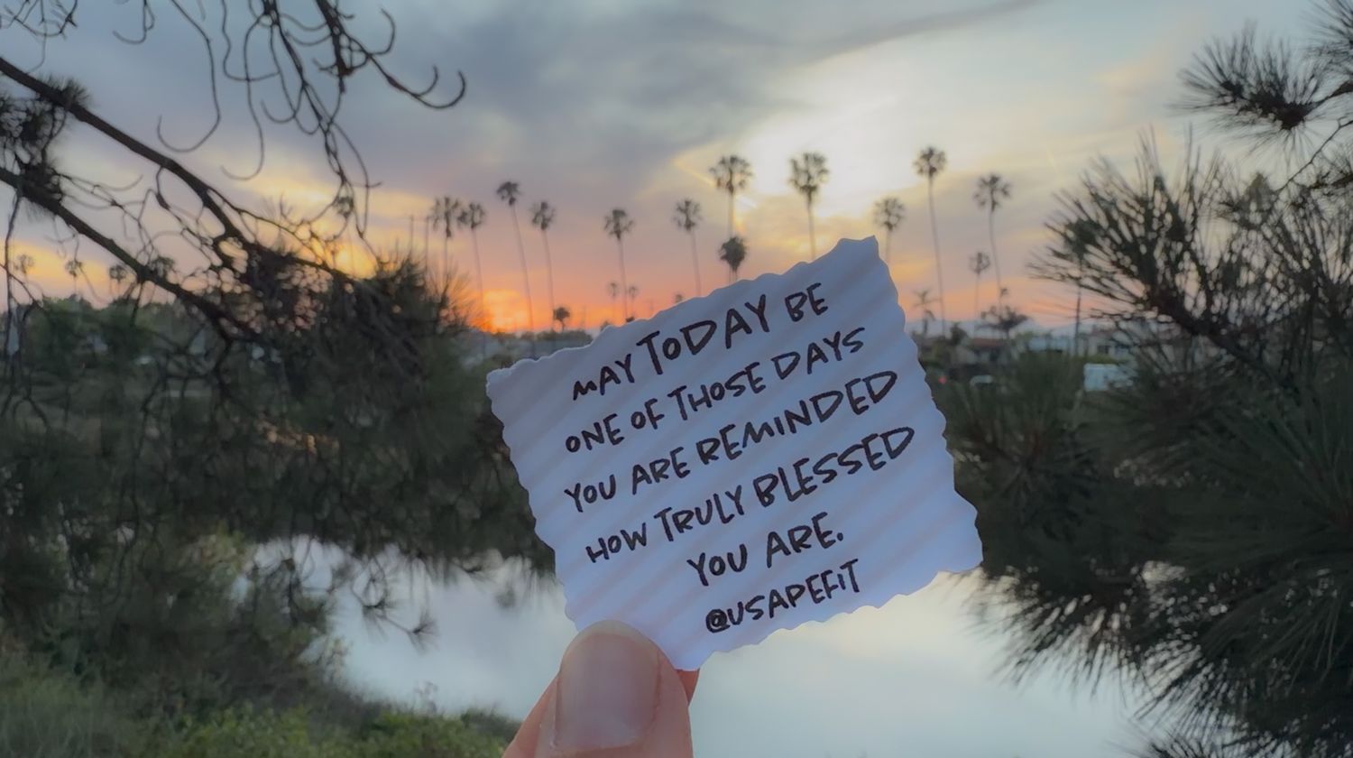 Hand holding motivational note at sunset with palm trees, reminding viewers how truly blessed they are.