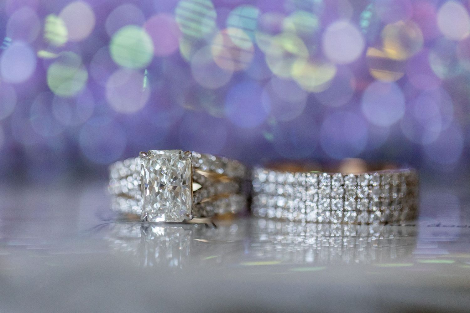 Macro shot of a radiant cut diamond engagement ring and diamond-encrusted wedding band with a purple and gold bokeh light background.