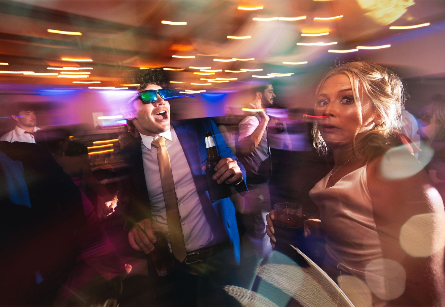 A joyful couple dancing energetically at a wedding reception, man in sunglasses holding beer, woman in elegant dress, colorful bokeh lights.