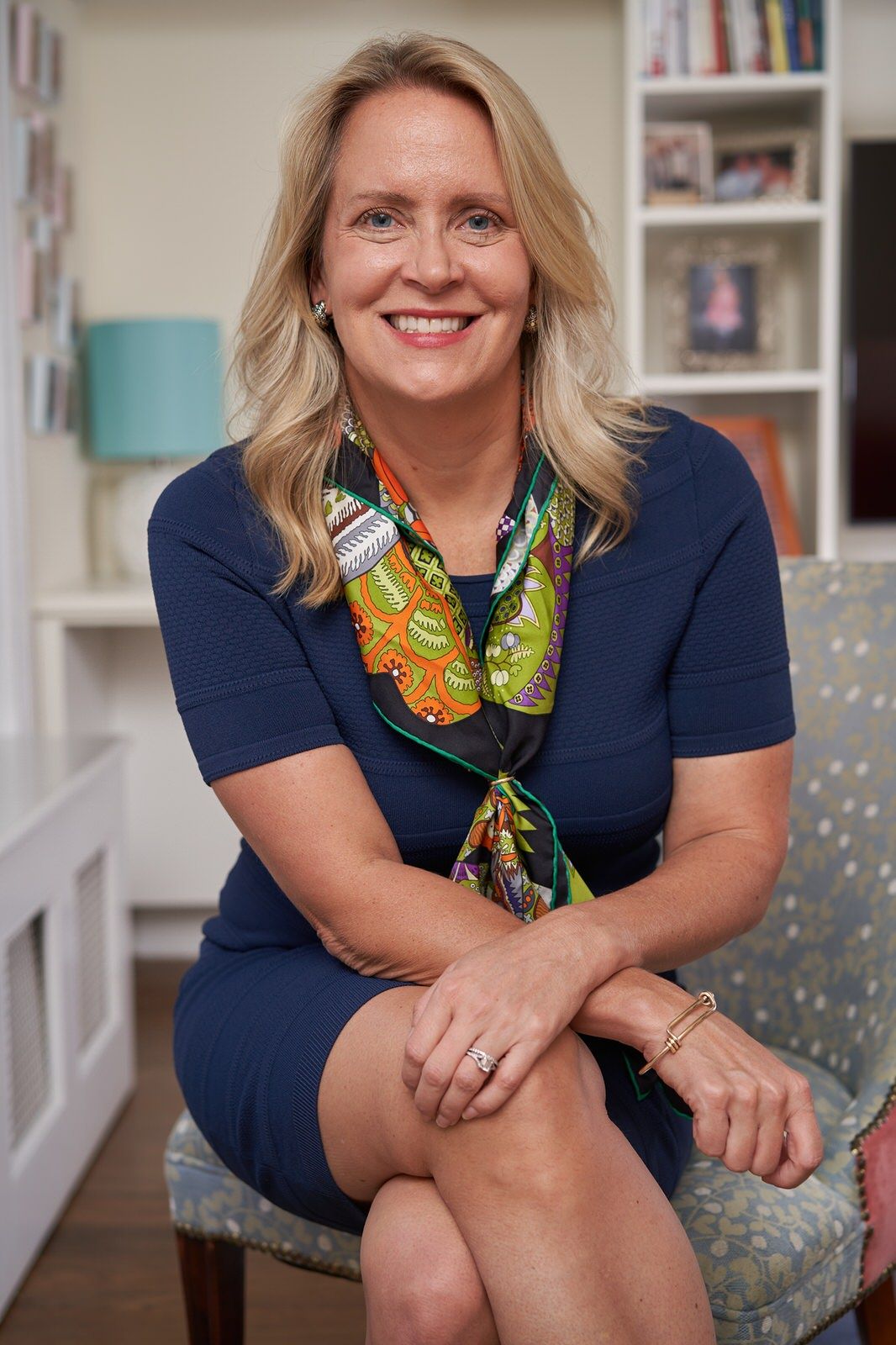 Smiling blonde woman in navy dress and colorful scarf sits cross-legged in chair in bright home office.