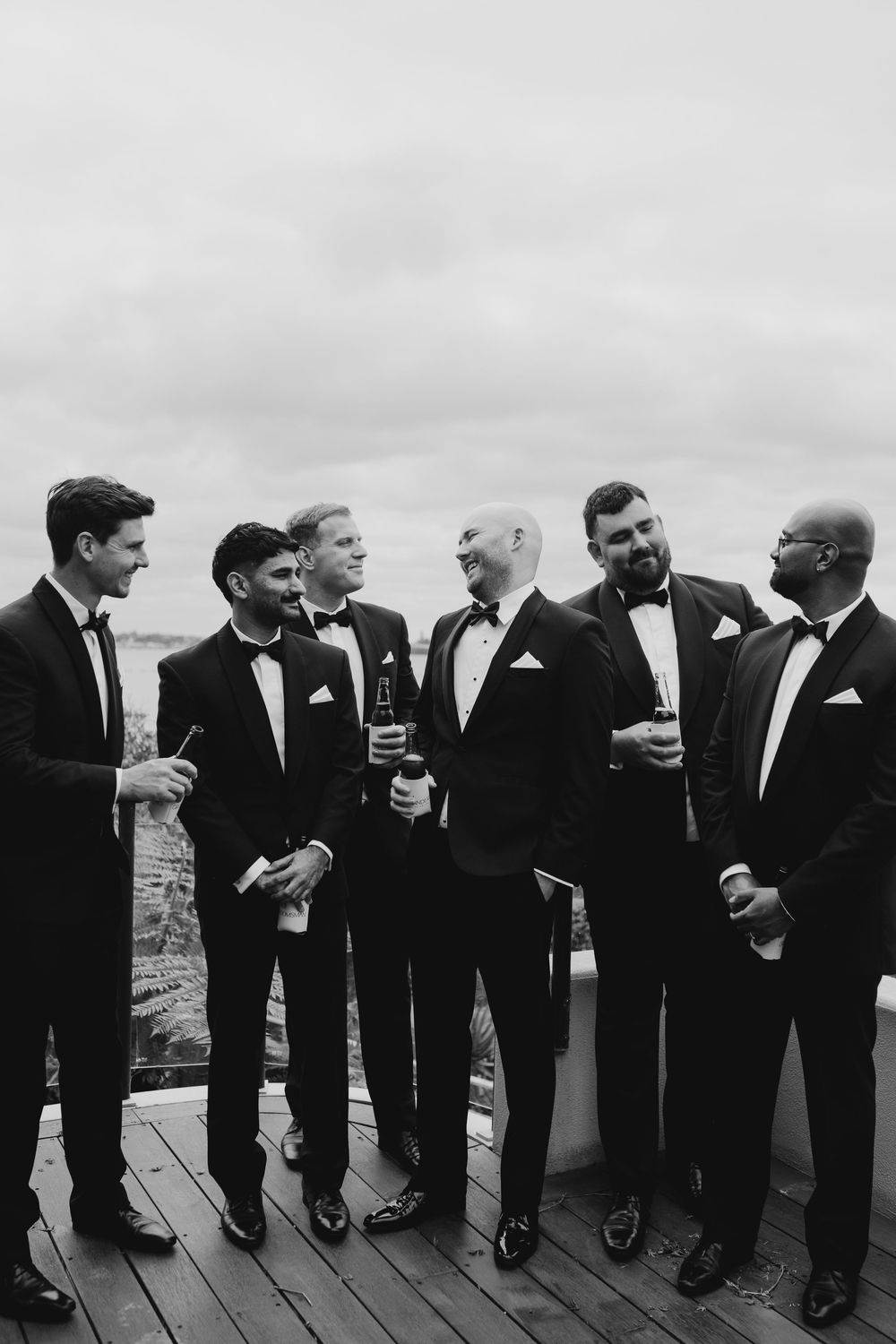 Black and white photo of groom and groomsmen in tuxedos holding drinks on rooftop.