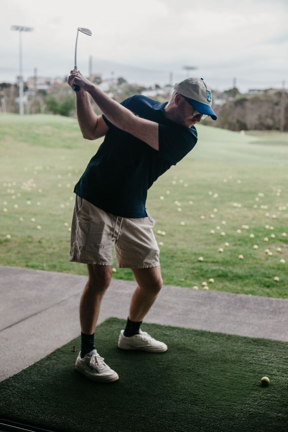 Man in cap and shorts practicing golf swing at driving range with yellow balls scattered on green.