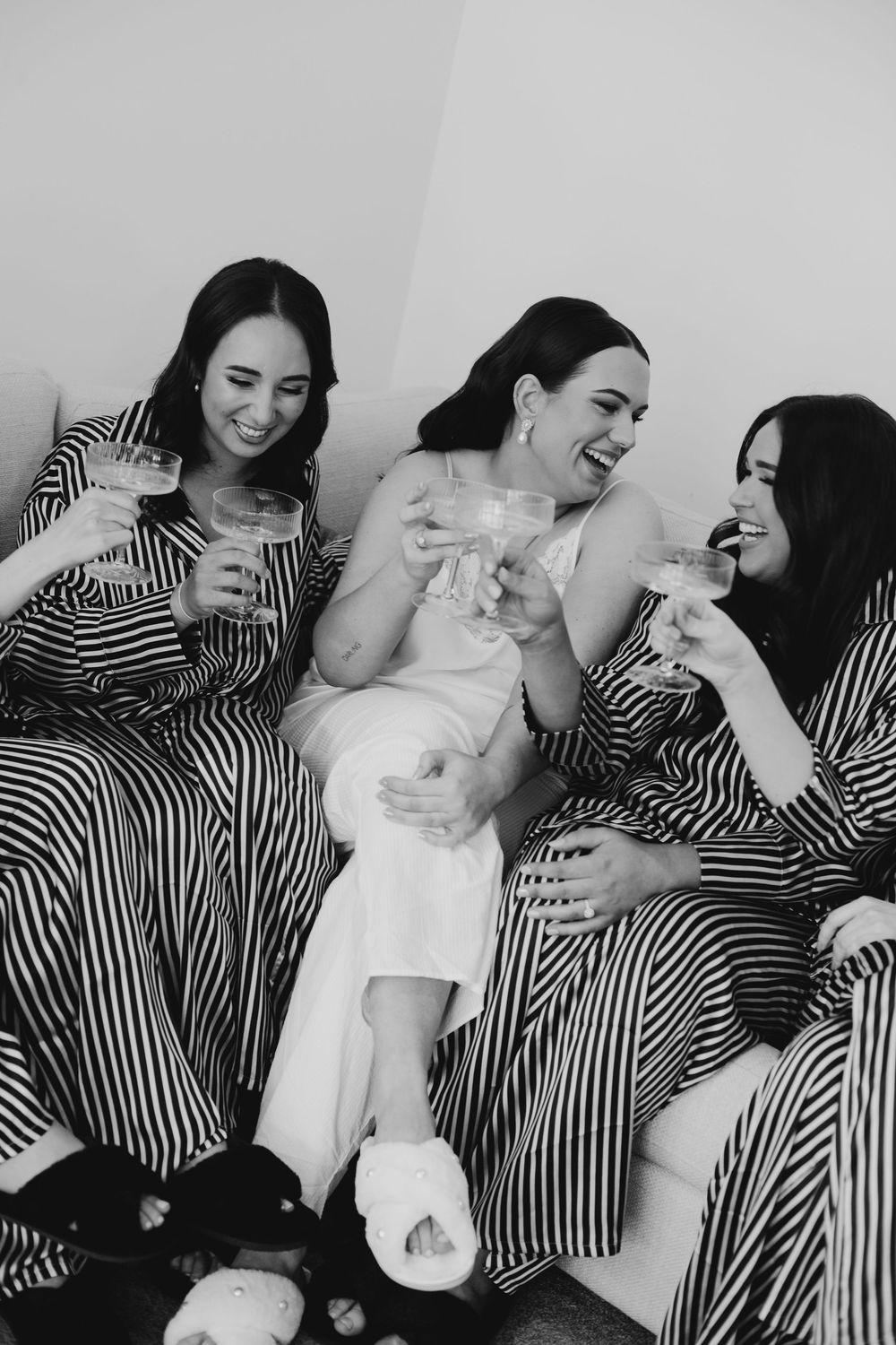 Black and white photo of bride and bridesmaids in stripe robes laughing with drinks.