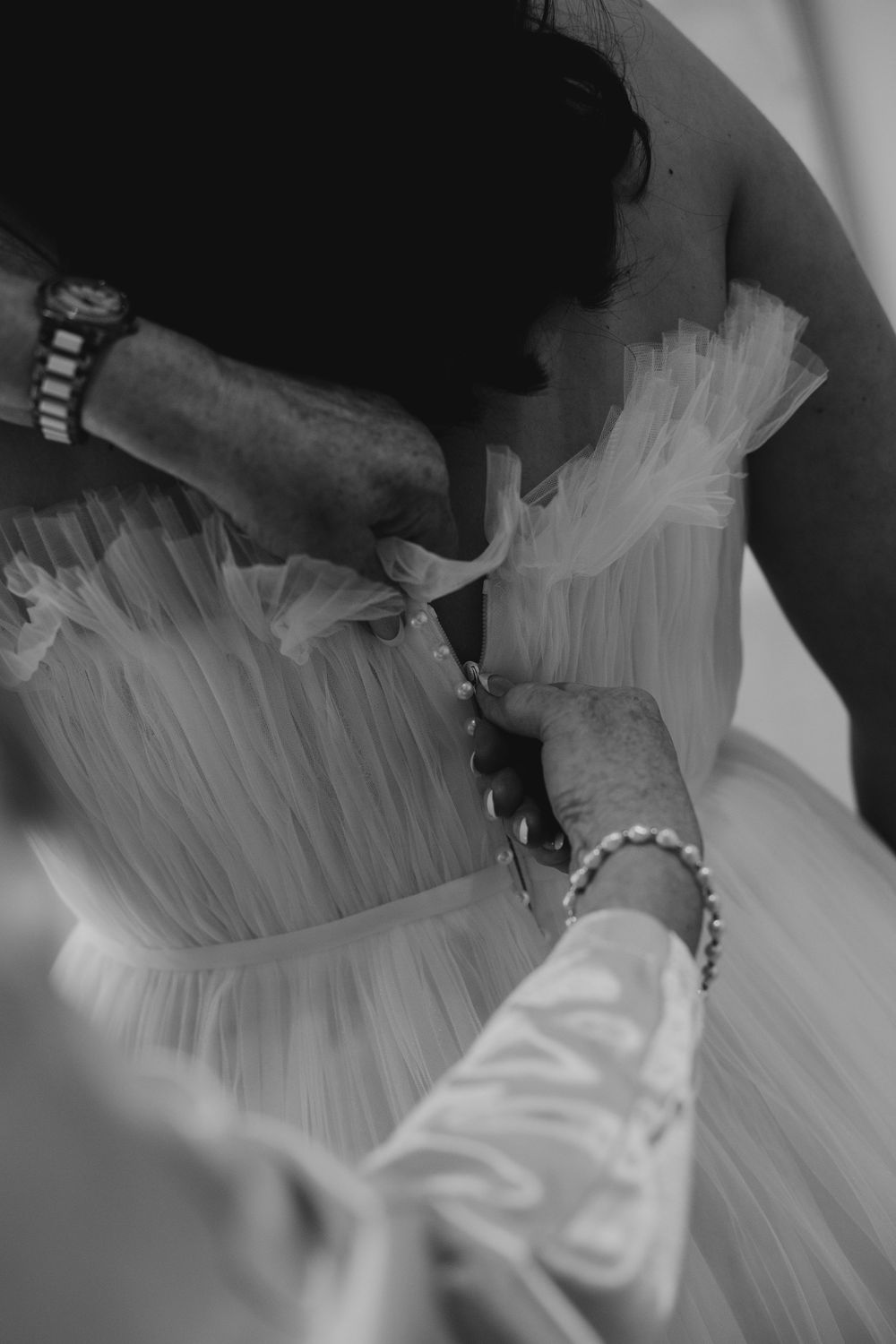 Black and white close-up of hands fastening buttons on back of white tulle wedding dress.