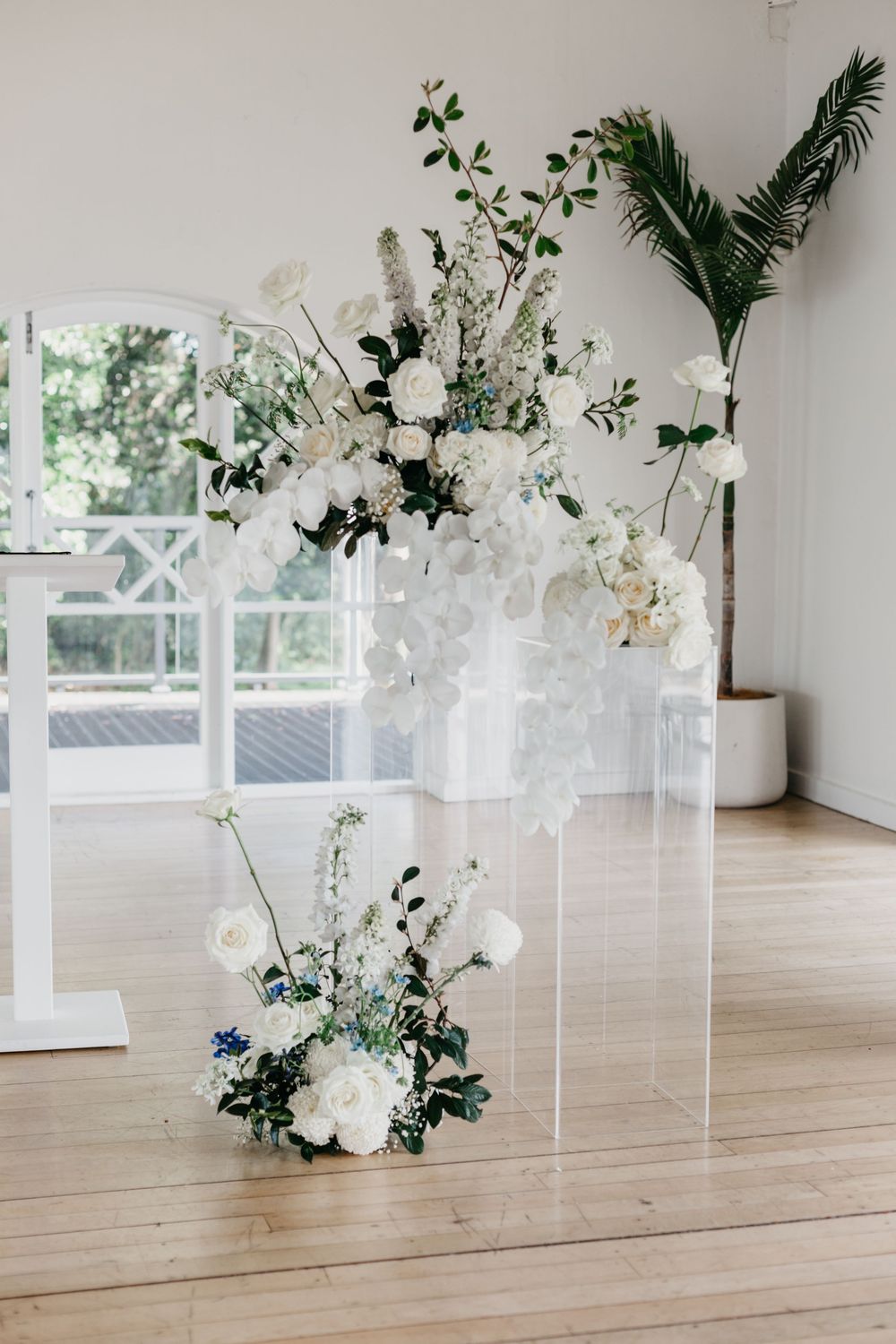 White floral wedding ceremony arrangement with roses and greenery on acrylic pedestal.