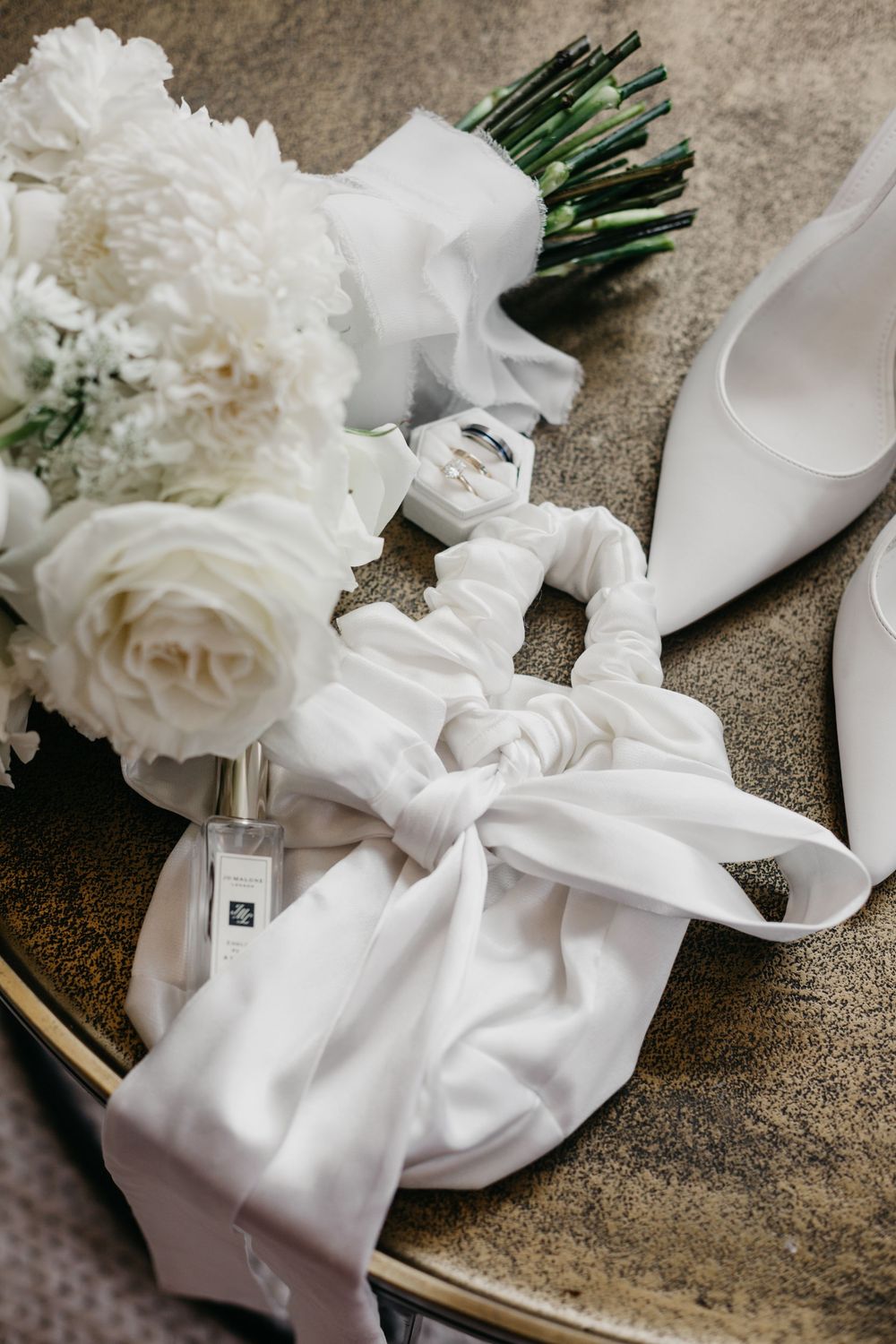 Bridal flat lay with white roses, perfume, ring box, hair scrunchie, and white heels.