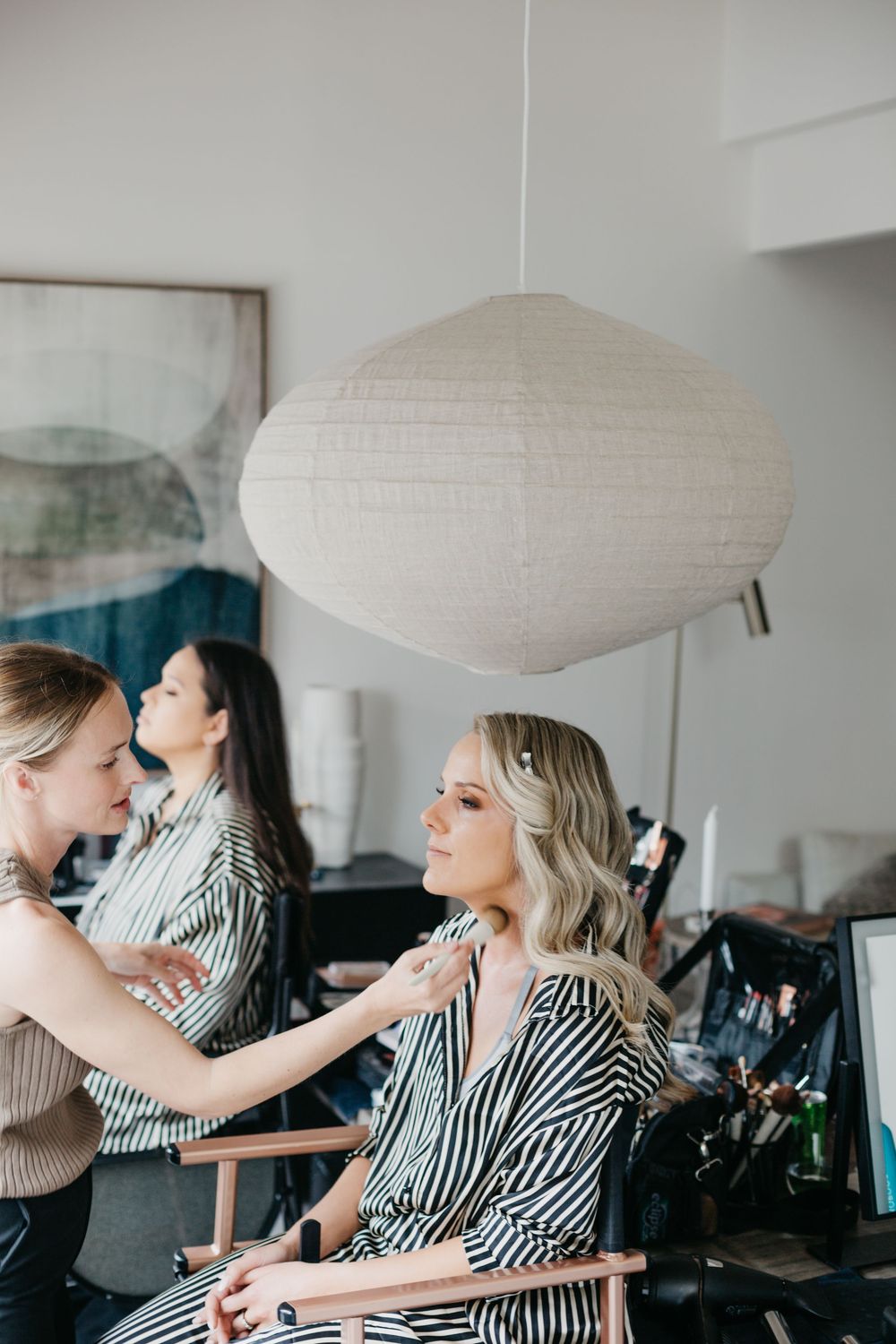 Blonde bride getting makeup applied by stylist in bright modern salon setting.