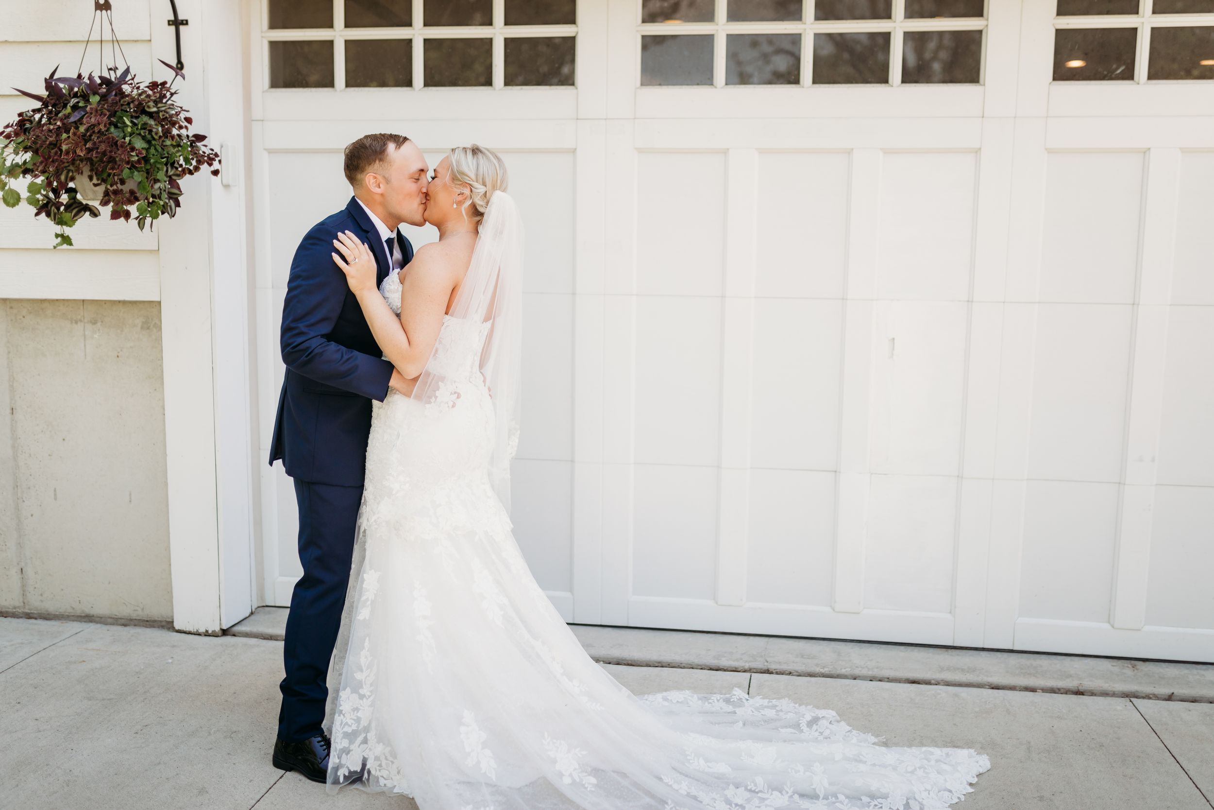 Bride and groom share a romantic kiss in front of a white garage door, bride in lace gown with veil, groom in navy suit.