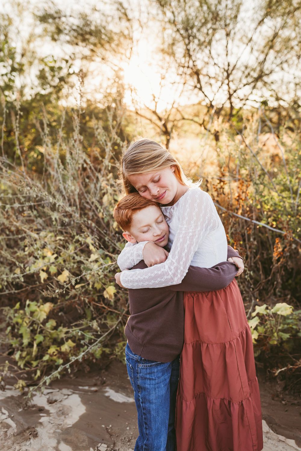 A girl and boy share a warm hug outdoors at golden hour, surrounded by desert shrubs.