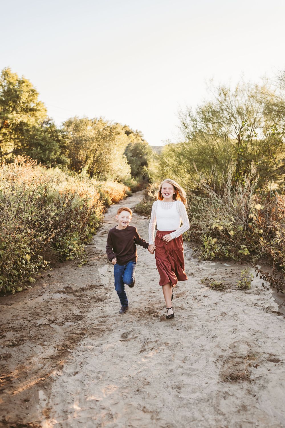 Two happy children run hand-in-hand down a sandy trail surrounded by lush greenery on a sunny day.