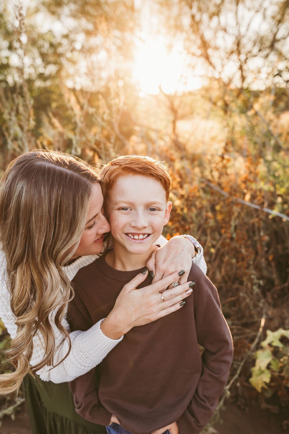 A mother hugs her red-haired son from behind, smiling outdoors in golden sunset light among autumn foliage.