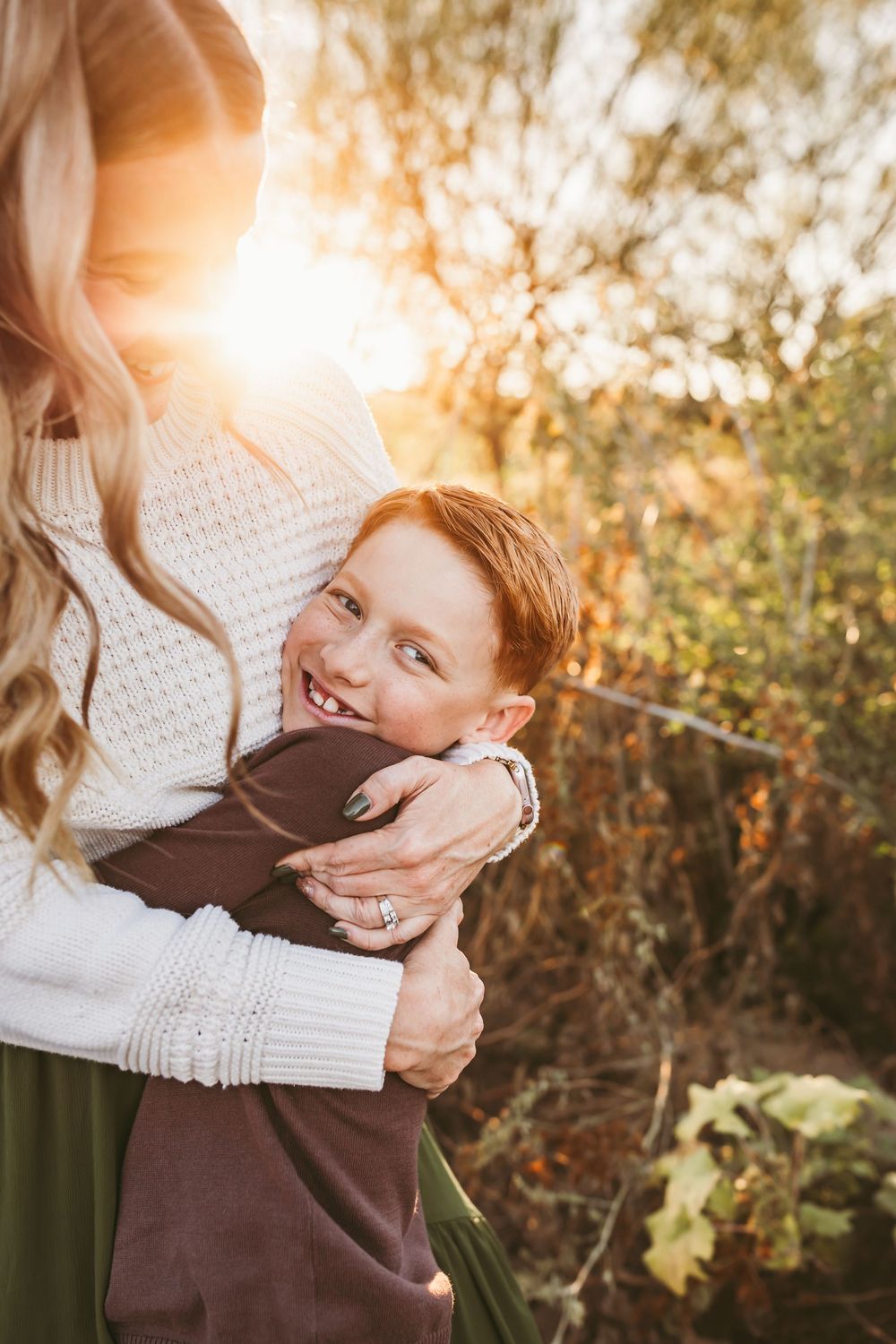 A red-haired boy smiles while being hugged by a woman in a white sweater outdoors in golden autumn sunlight.