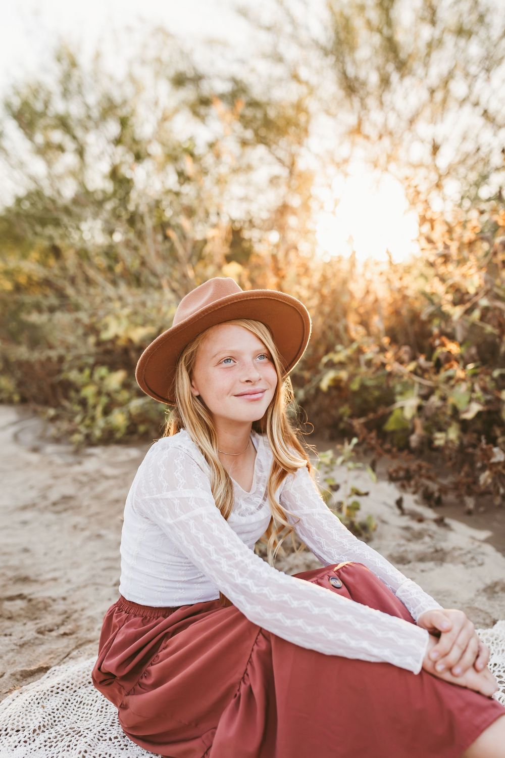 Young girl in brown hat and white lace top sits on sandy beach path at golden hour sunset.