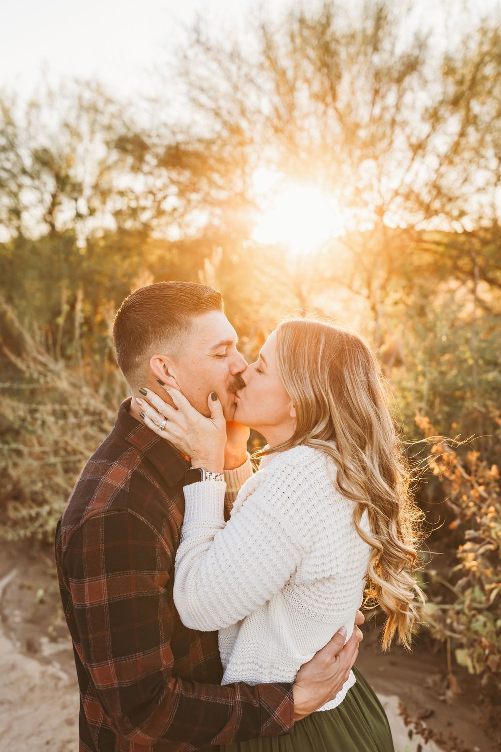 A couple shares a romantic kiss at golden hour, embraced in a sun-drenched desert landscape.