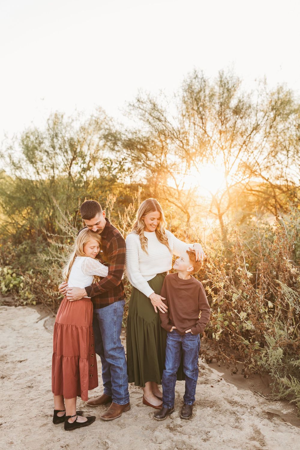 A happy family of four poses together outdoors in warm sunset light, surrounded by natural desert greenery.