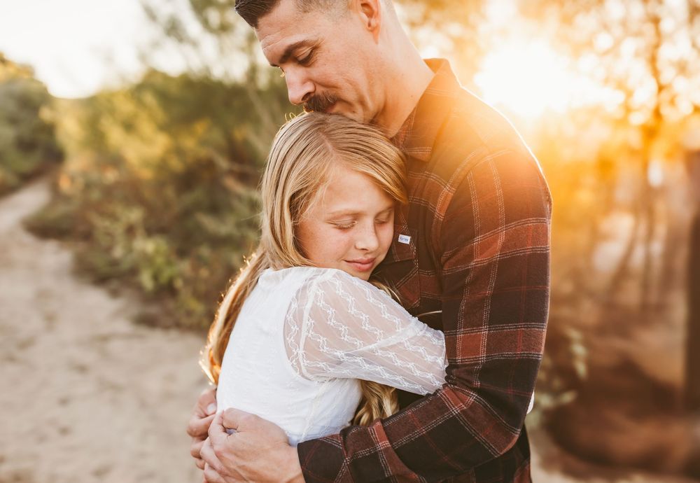A father and daughter share a warm embrace outdoors at golden hour, surrounded by trees and soft sunlight.