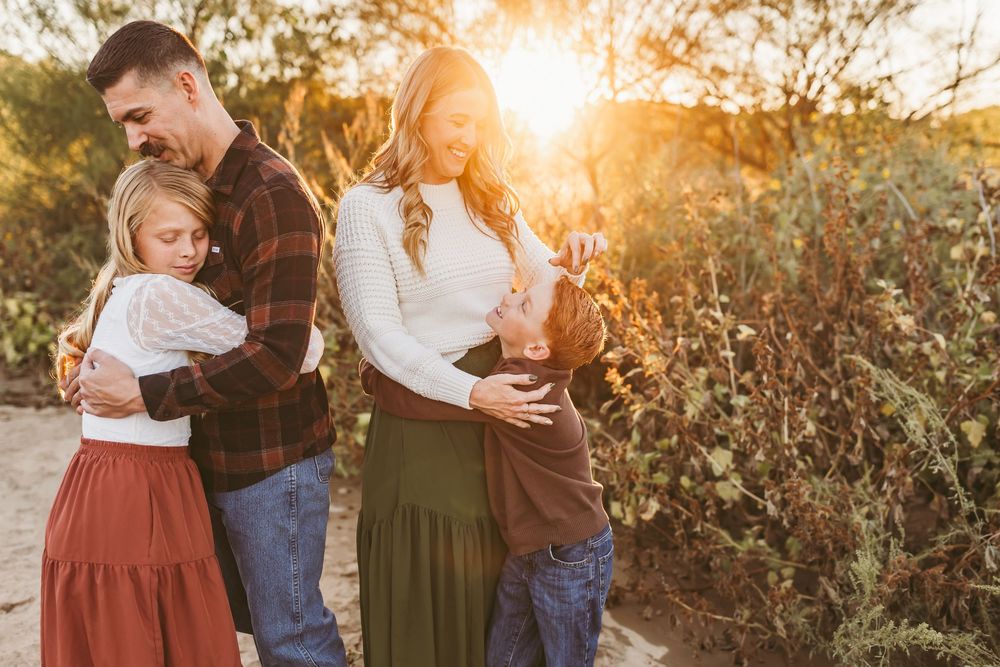 Family of four embracing outdoors at golden hour sunset, parents and two children smiling in autumn-toned outfits.