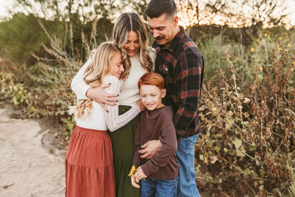 A happy family of four embraces outdoors in fall, with two children hugging their parents among autumn foliage.