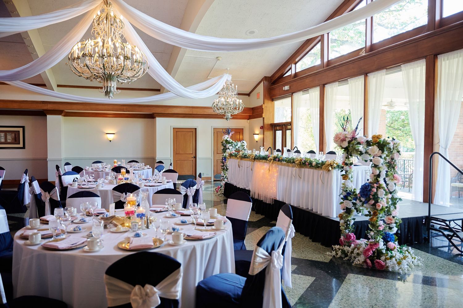 Elegant wedding reception hall with white draped ceilings, chandelier, floral arch, and round tables with black and white chair covers.